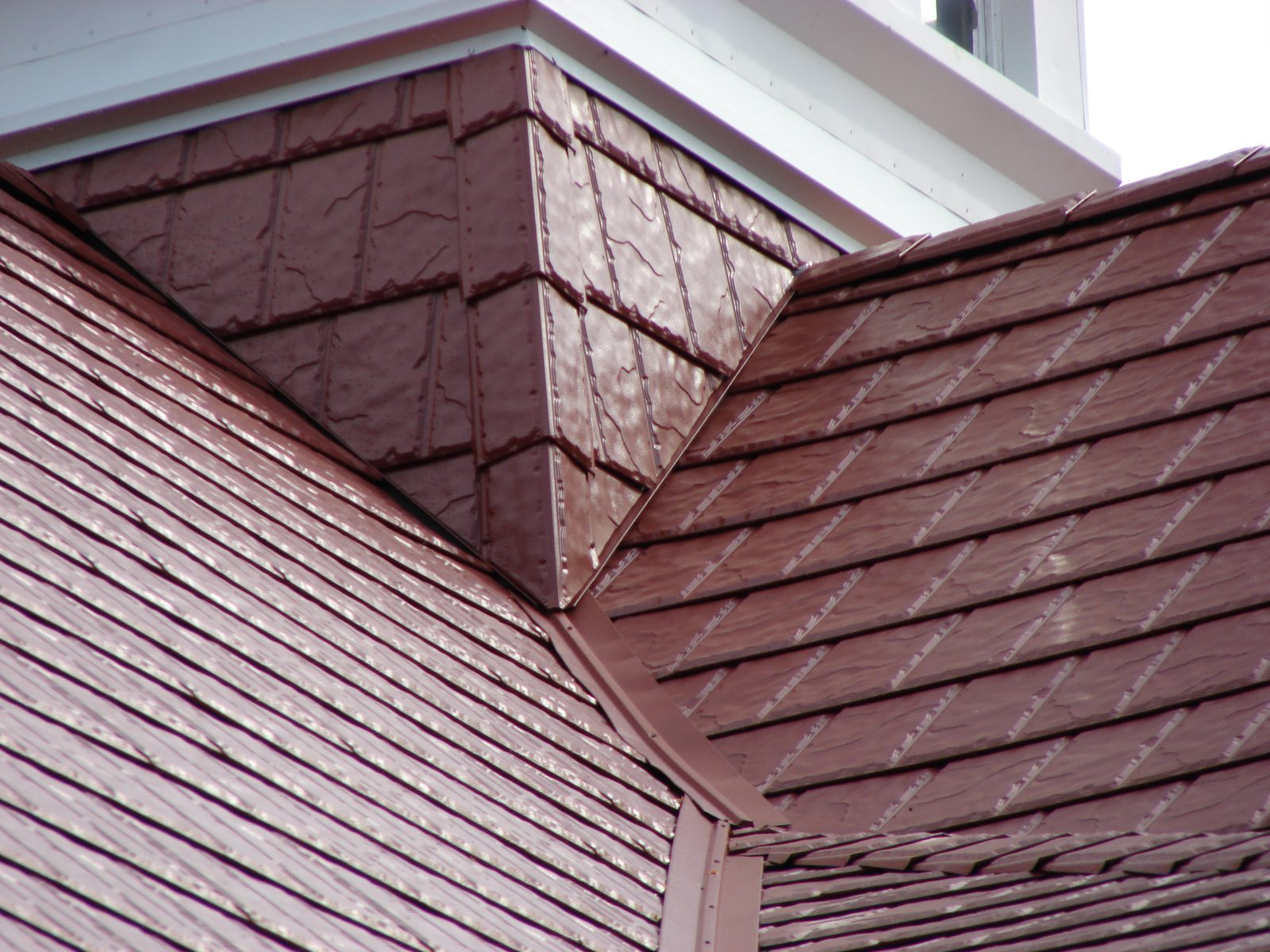 Dark red steel shake panels meet at a ridge and valley beneath white trim and a soffit return.