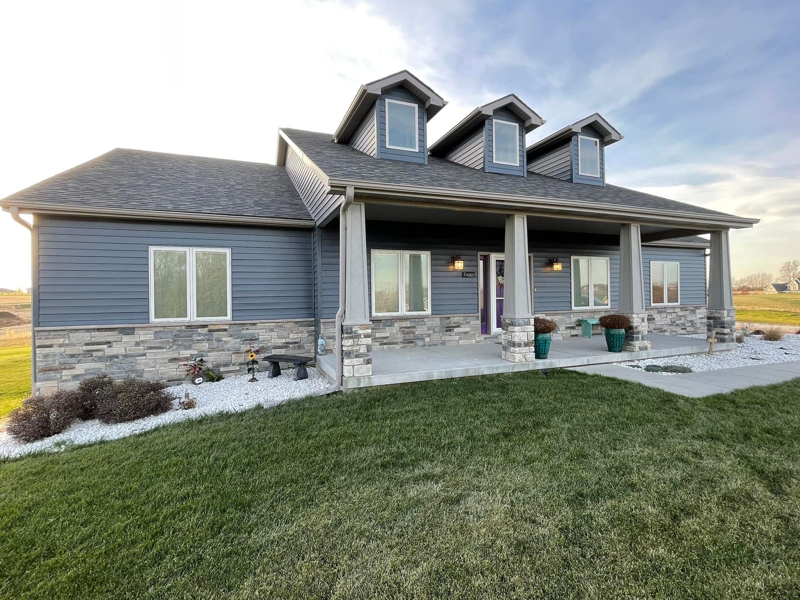 Blue-gray siding and a stone wainscot on a ranch home with three dormers and a covered porch.