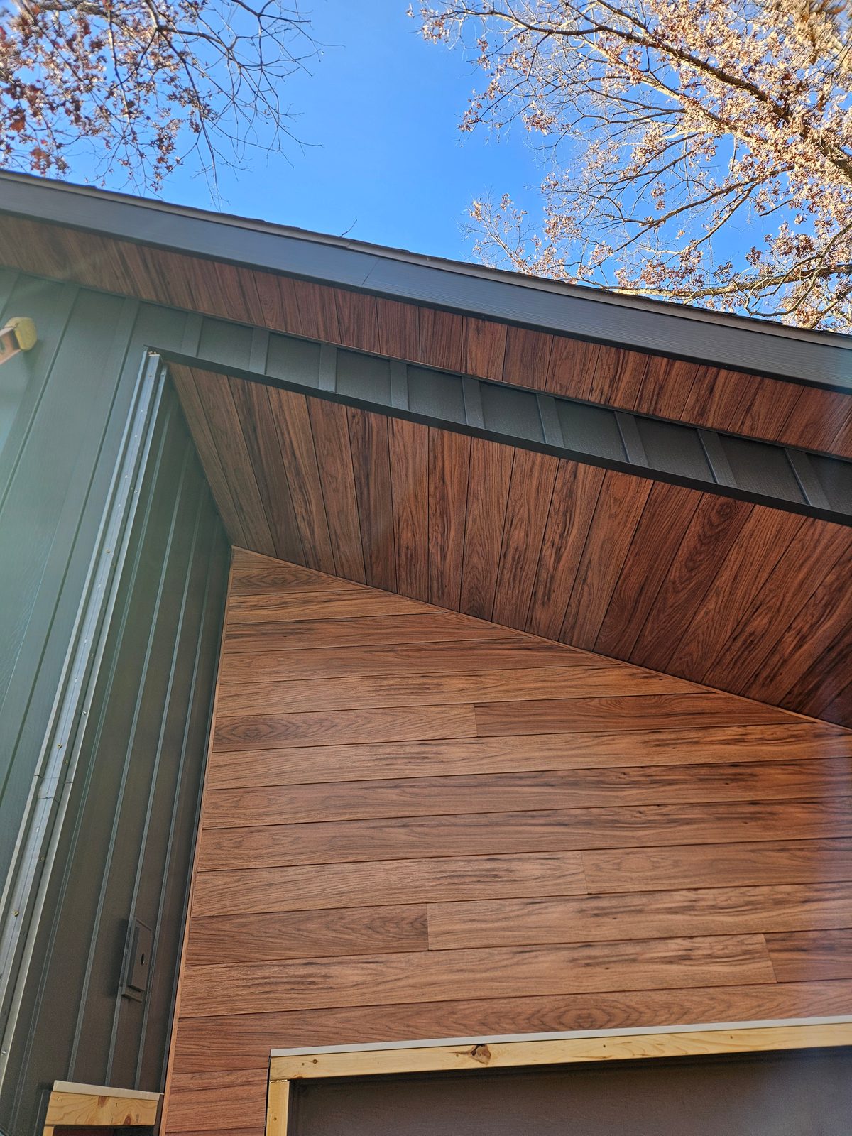 Wood-look steel siding covers a gable end and its soffit next to sage green vertical panels.