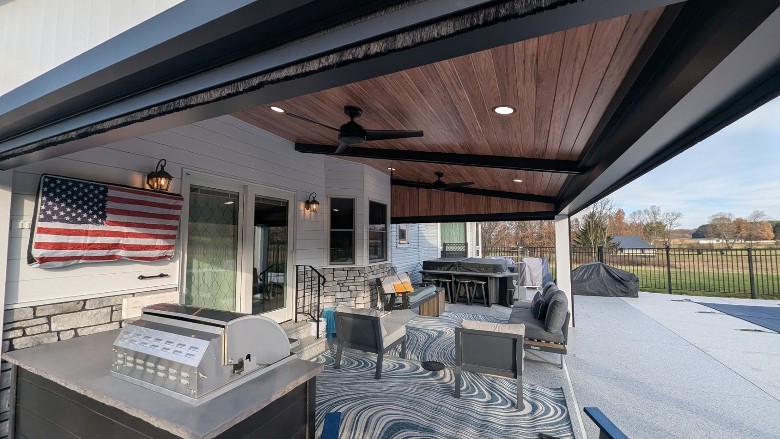 Wide view of a covered porch with wood-look steel ceiling panels, a grill station and seating.