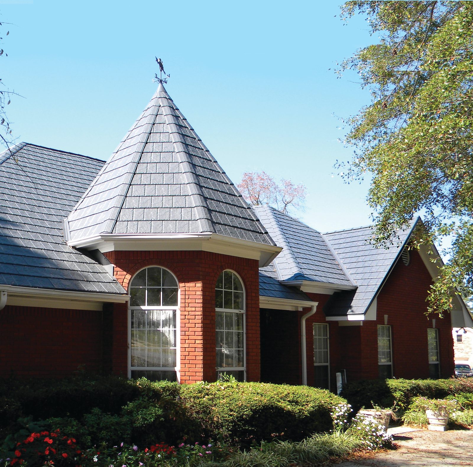 Gray steel shake roofing covers a conical turret and steep gables on a red brick home.