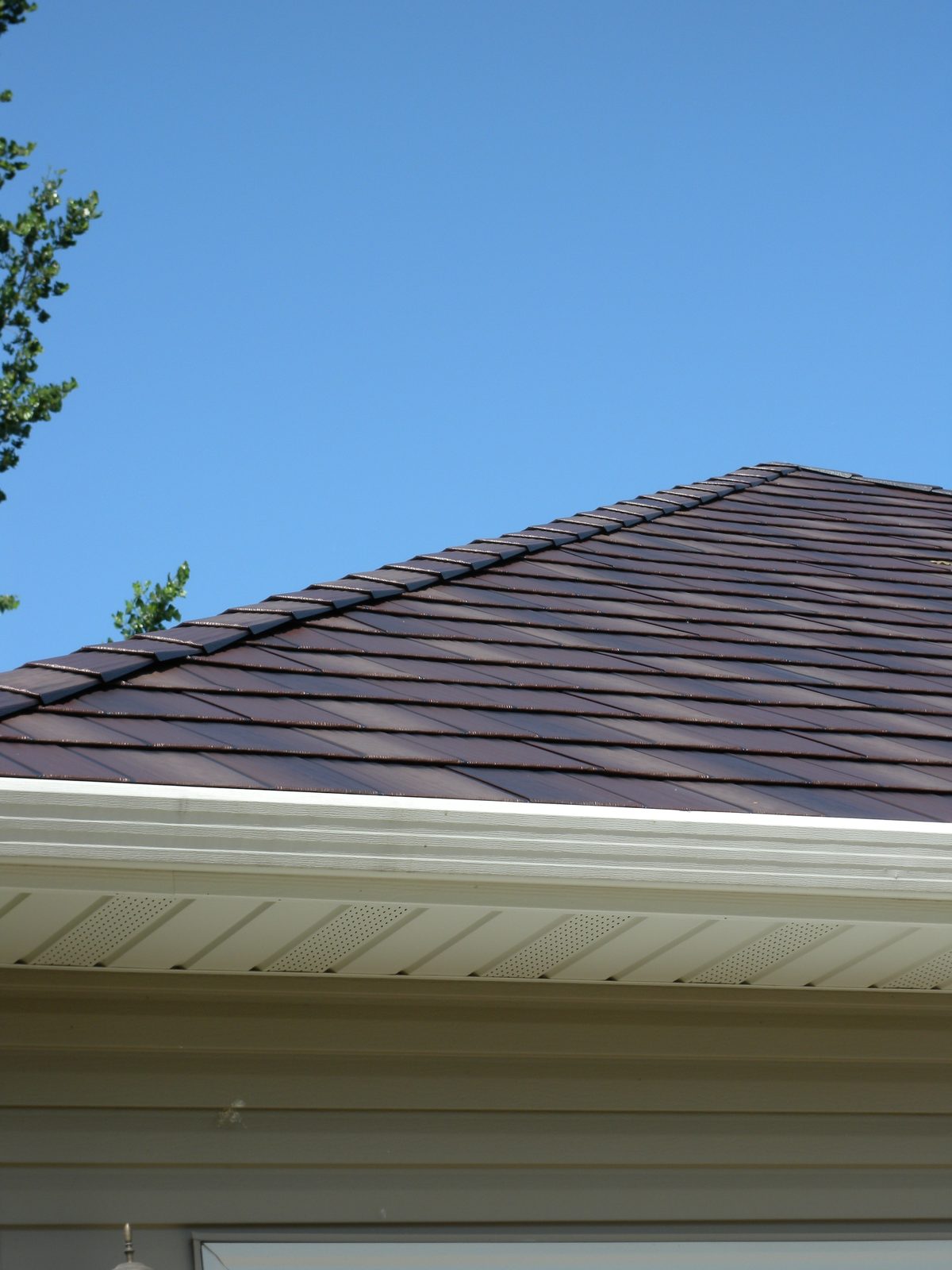 Dark brown steel shake roofing meets a white gutter and vented soffit against a clear blue sky.