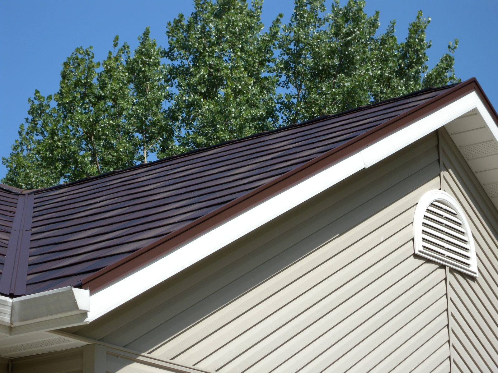 Brown steel roofing panels run down a gable slope beside tan lap siding and a louvered vent.