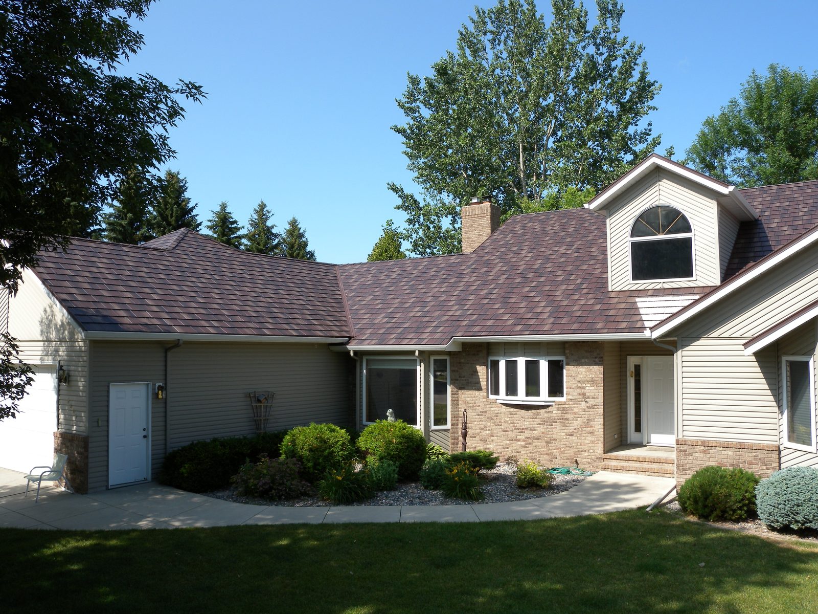 A tan two-story home wearing a new dark brown steel shake roof under a clear sky.
