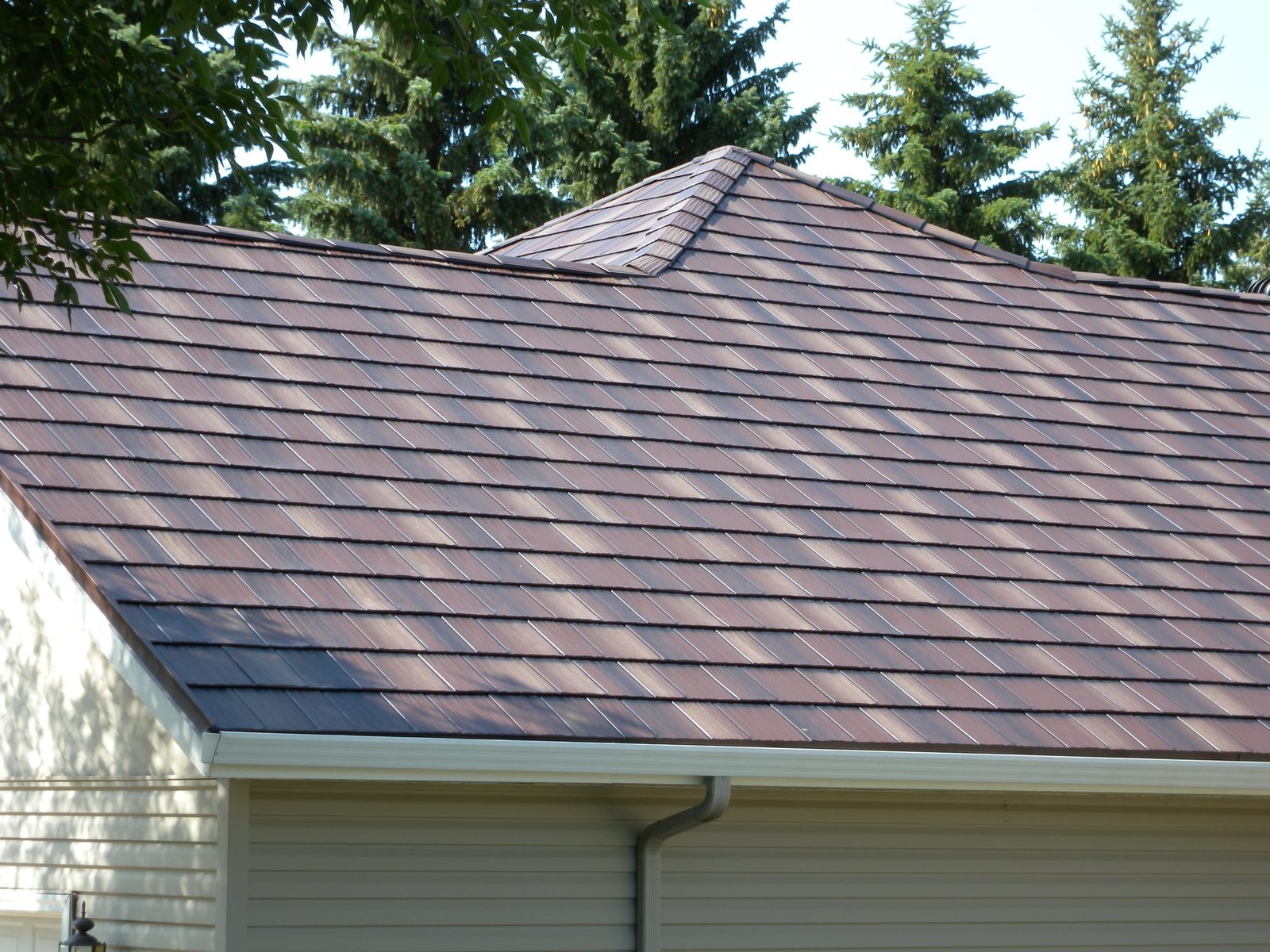 Brown steel shake roofing covers a hipped roof plane above a garage and tan lap siding.
