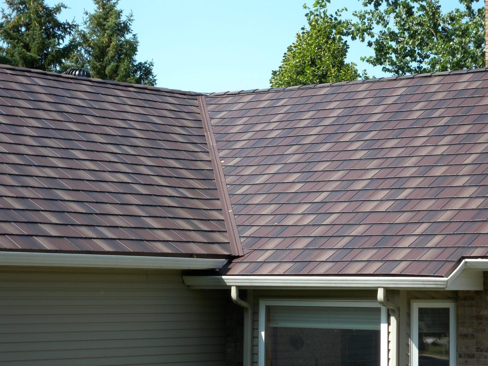 Two brown steel shake roof planes meet along a ridge above a bay window and white gutter.
