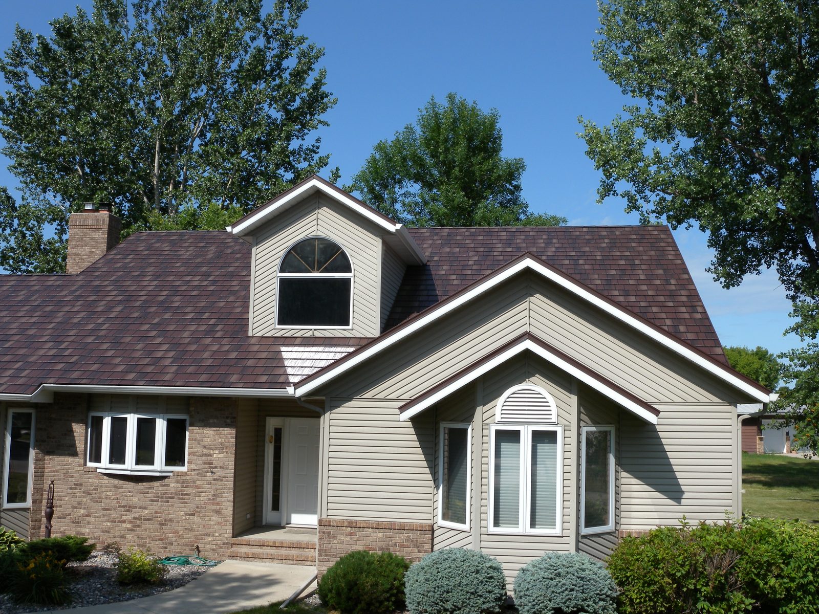 A tan home with an arched dormer window and a new brown steel shake roof against a blue sky.