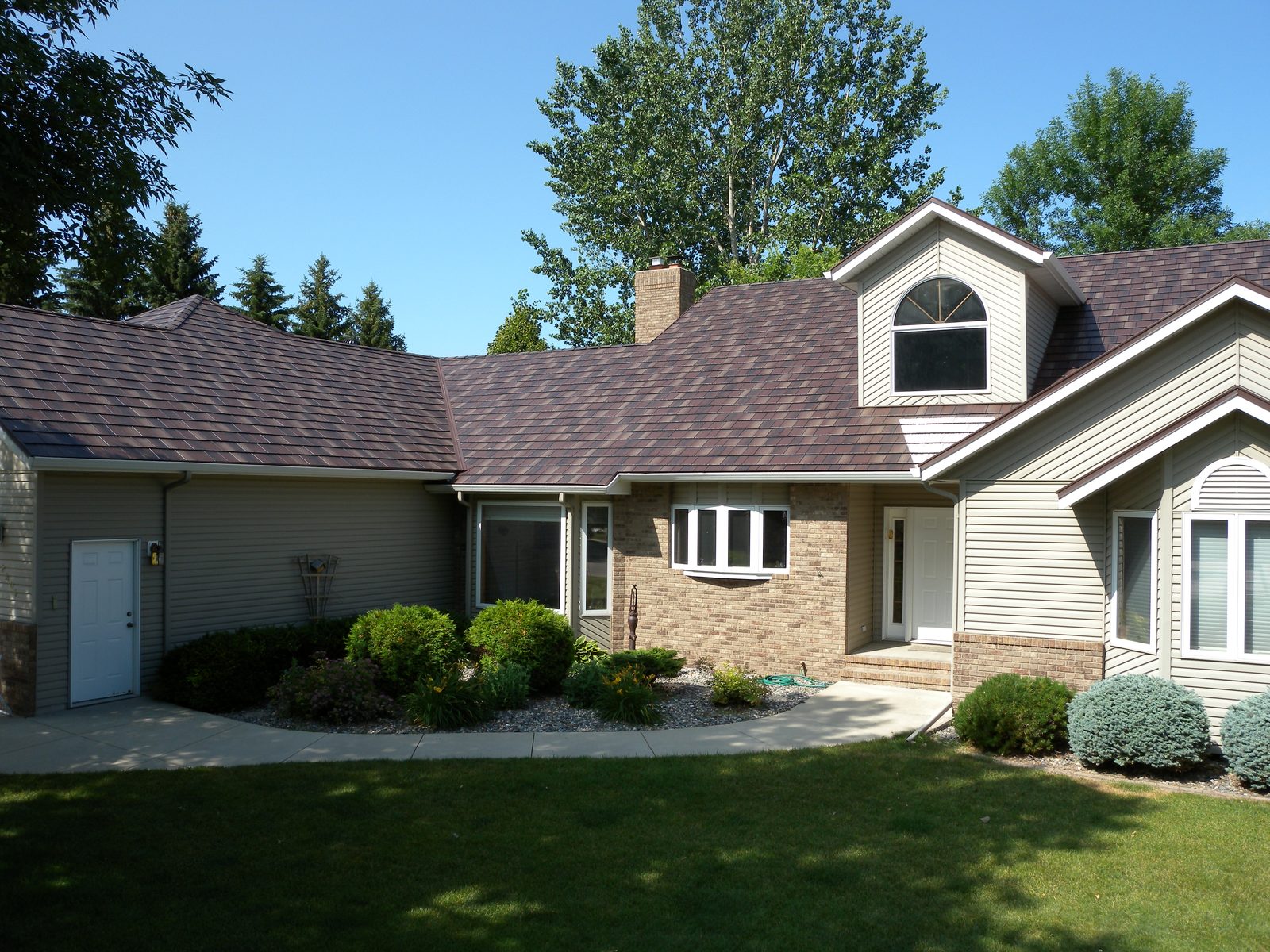A tan two-story home with brown steel shake roofing, viewed from the edge of the driveway.