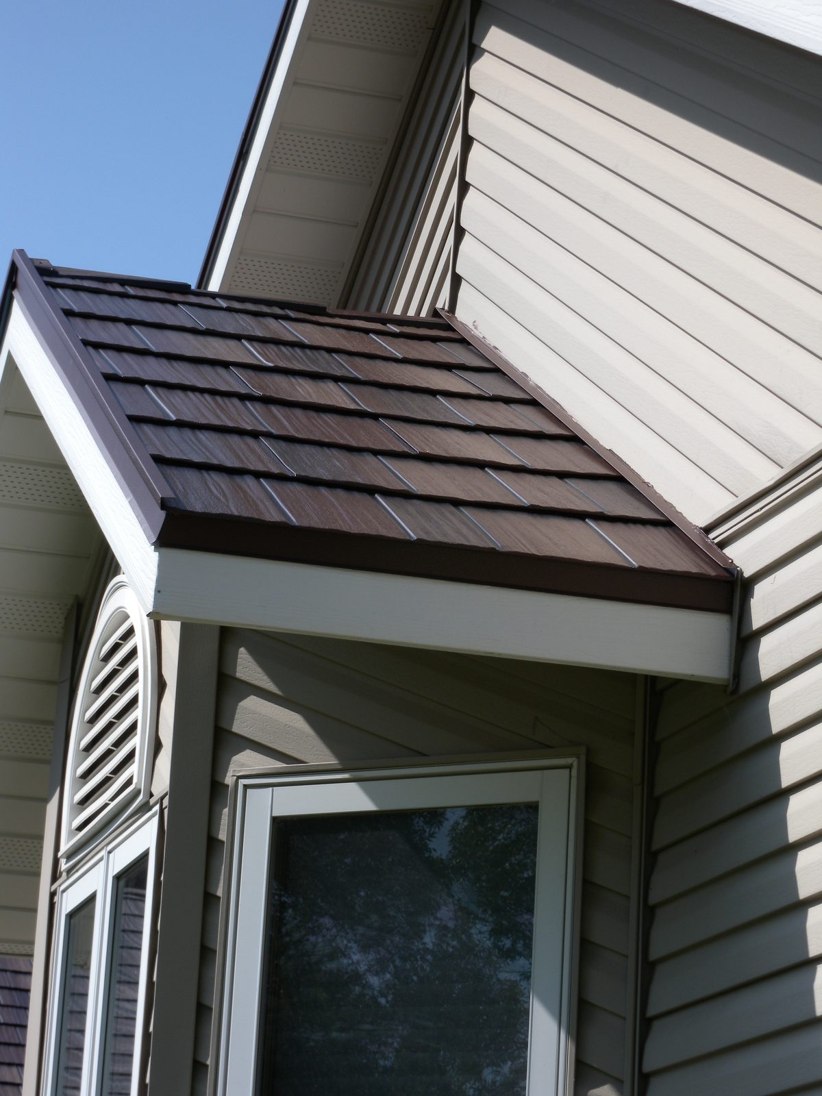 A small brown steel shake roof caps a bay window projection below tan lap siding.