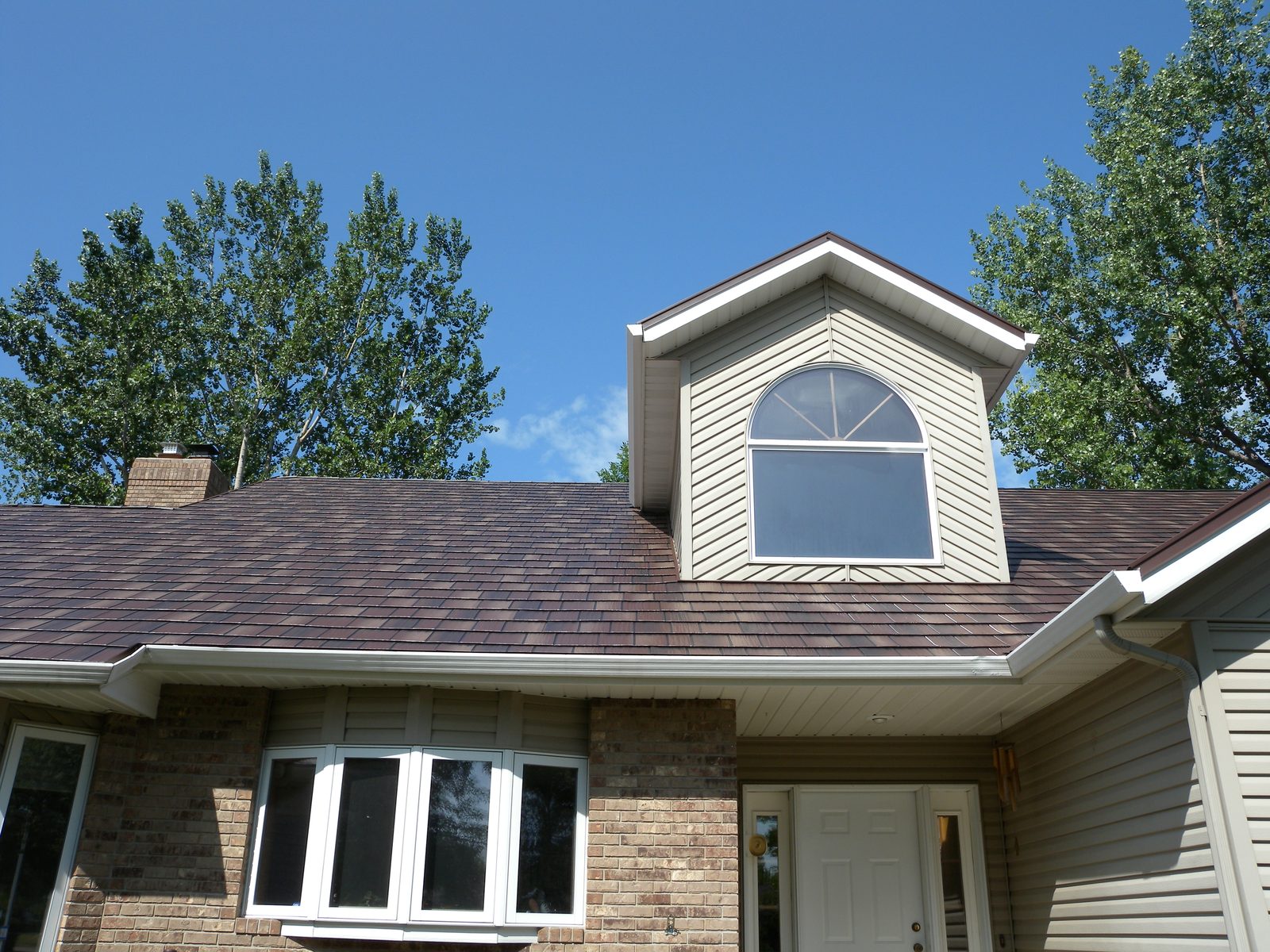 An arched dormer window rises above a brown steel shake roof on a tan brick home.