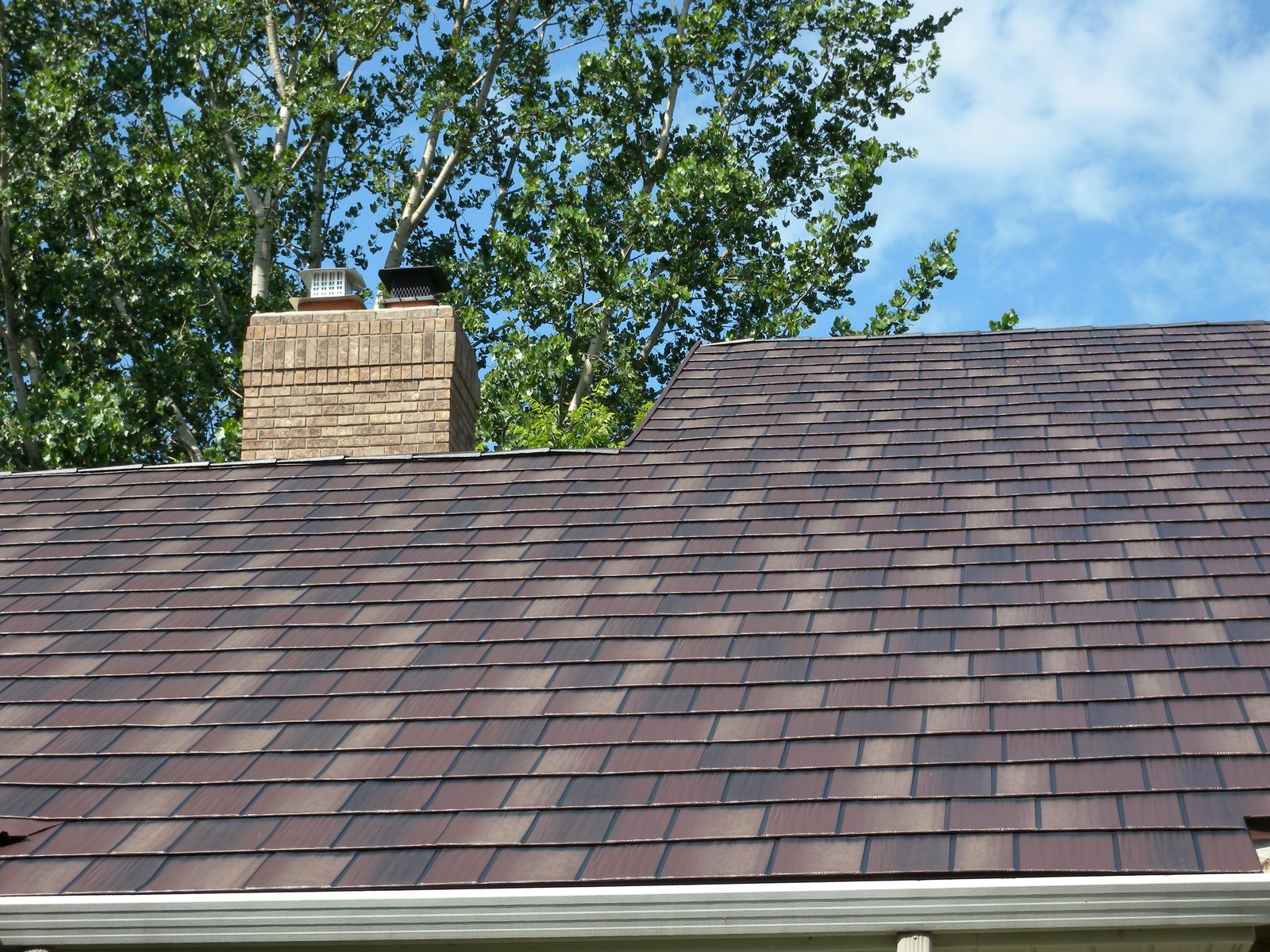 Brown steel shakes surround a tan brick chimney below leafy branches and open sky.