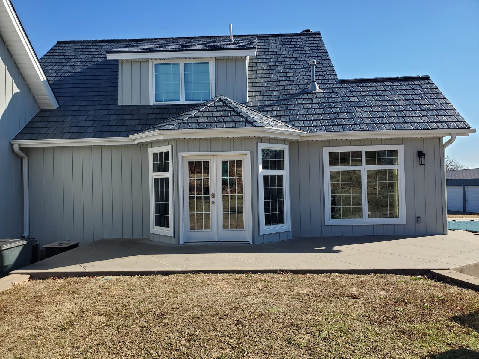 Rear elevation of a gray farmhouse with steel shake roofing above a bayed sunroom and patio.