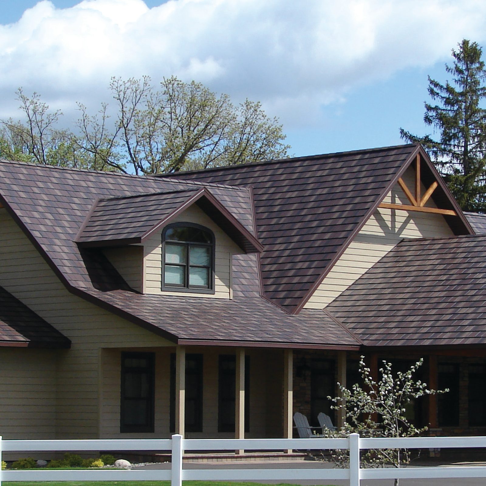 Plum-brown steel shake roofing spans intersecting gables above tan siding and a timber gable truss.