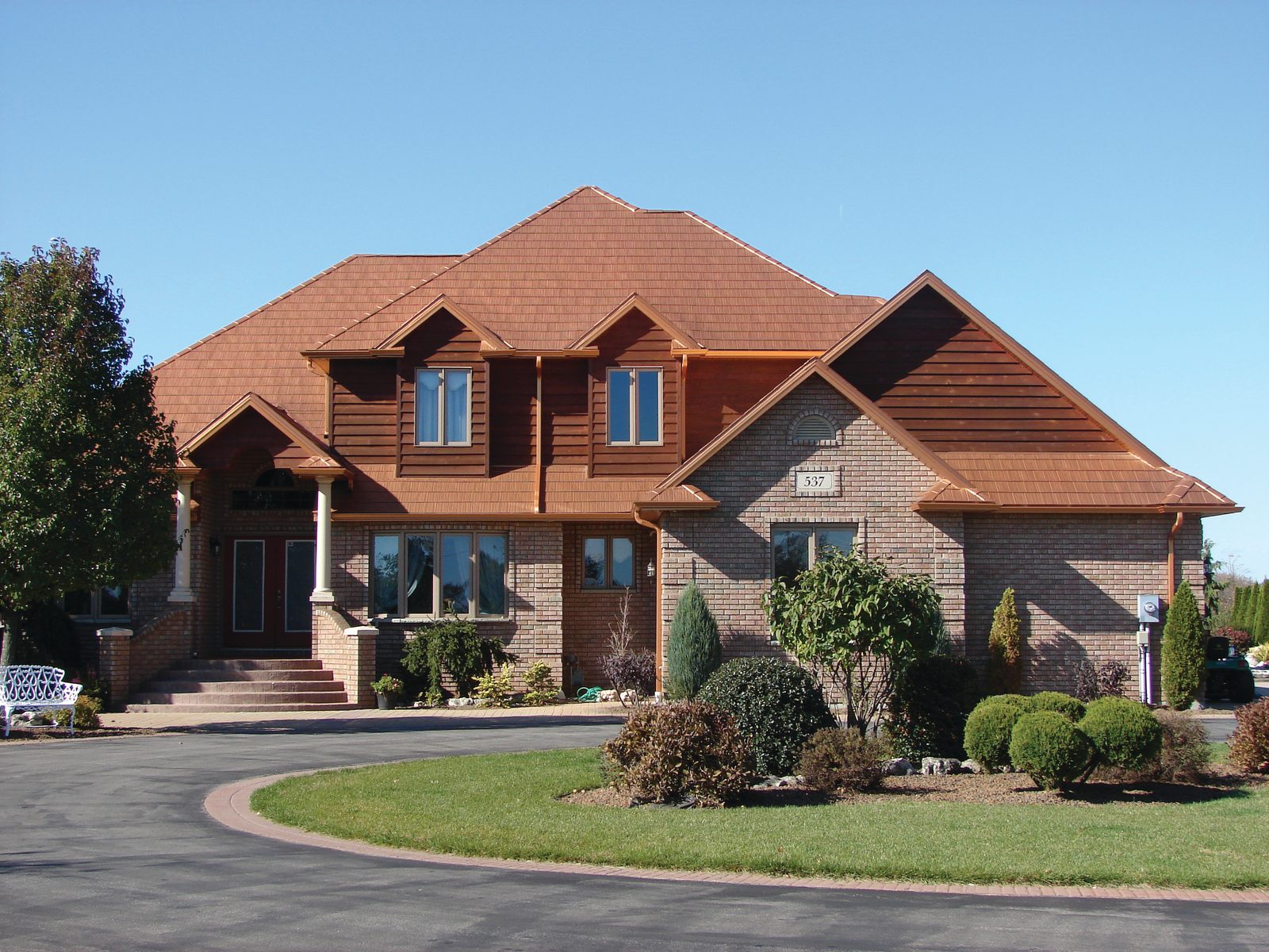 Copper-toned steel shake roofing covers the multiple gables of a large brick and log-accent home.