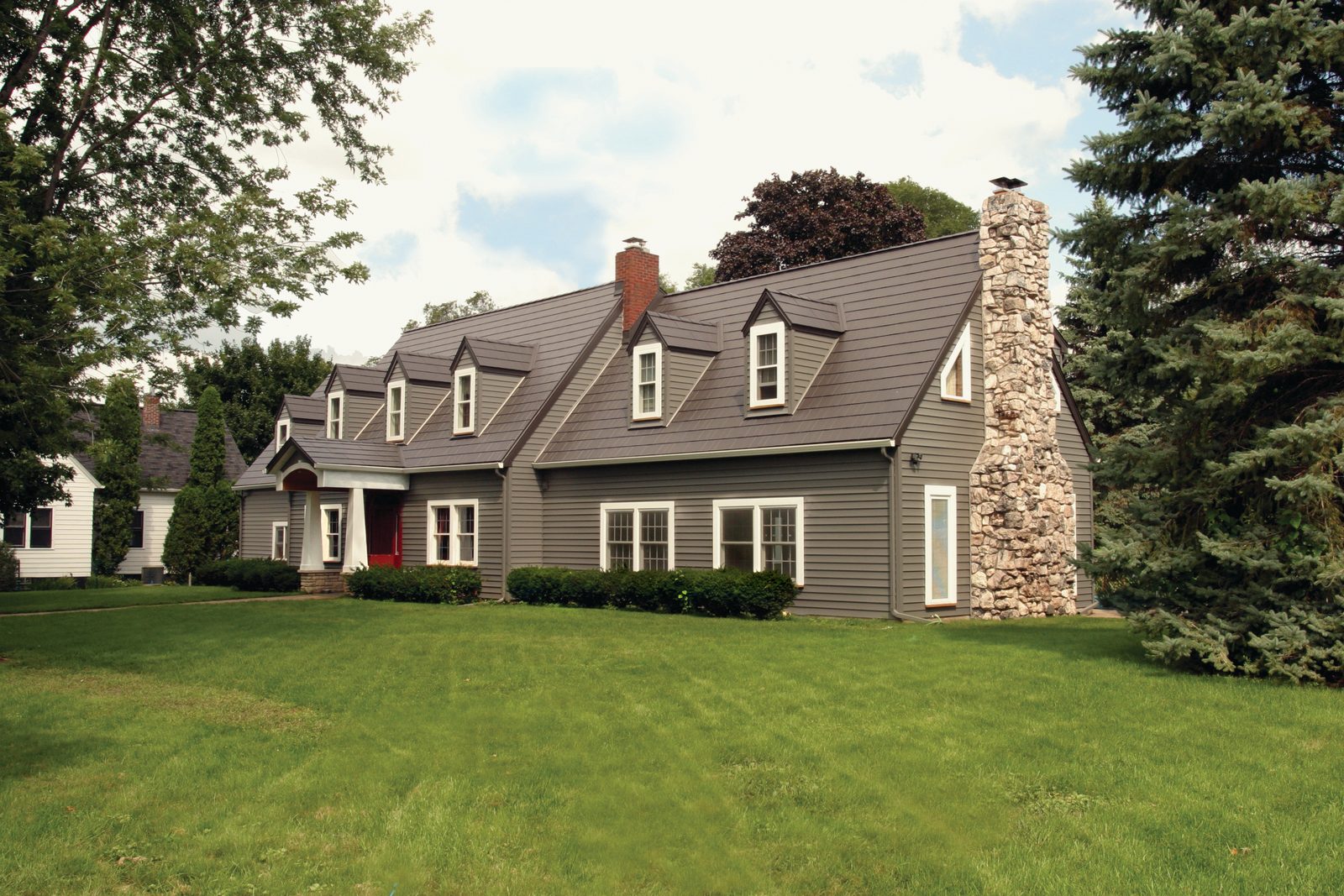 Gray steel shake roofing covers a gambrel roof with four dormers beside a tall fieldstone chimney.