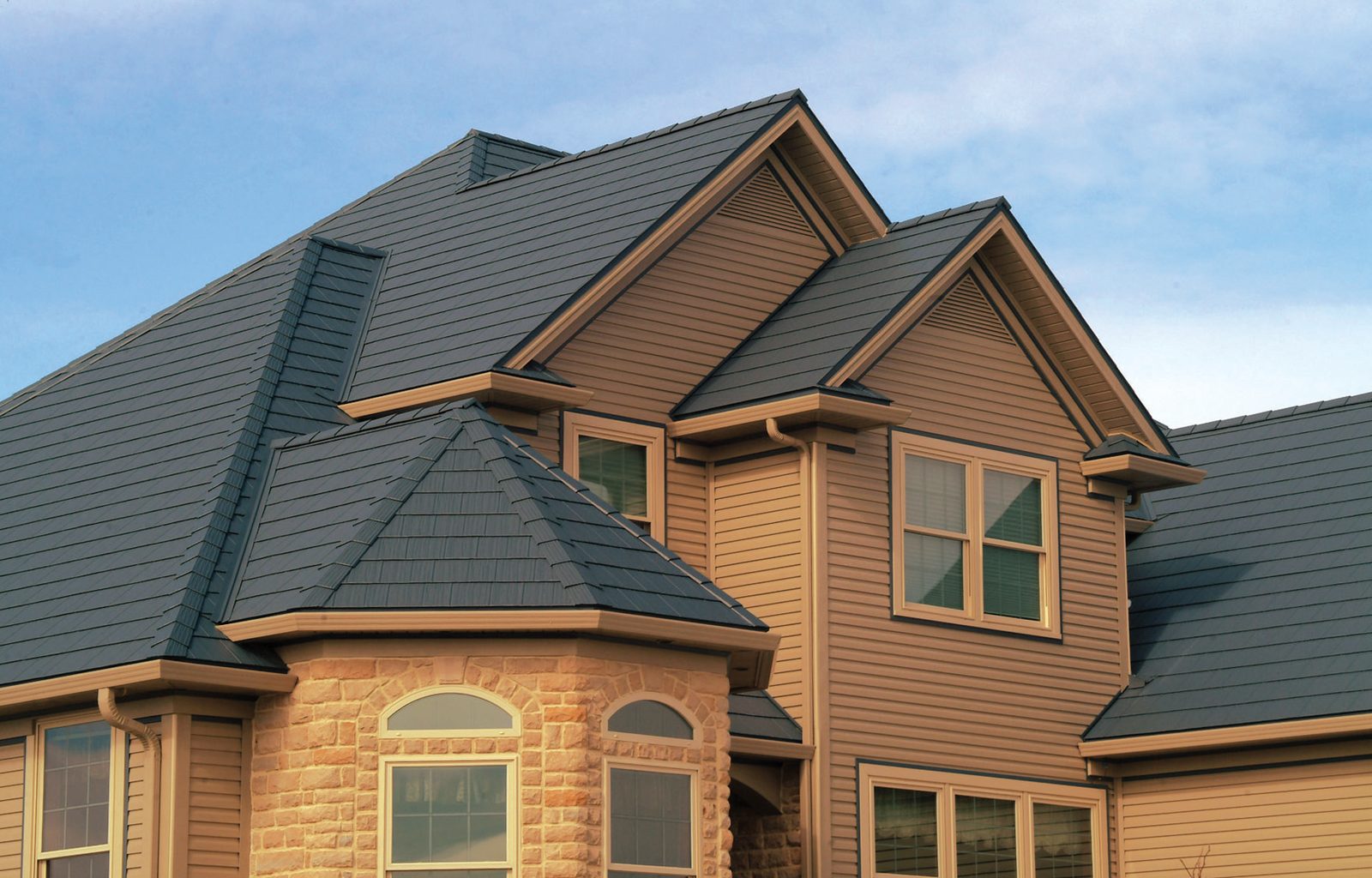 Dark blue steel shake roofing covers the hips and gables of a large tan two-story home.