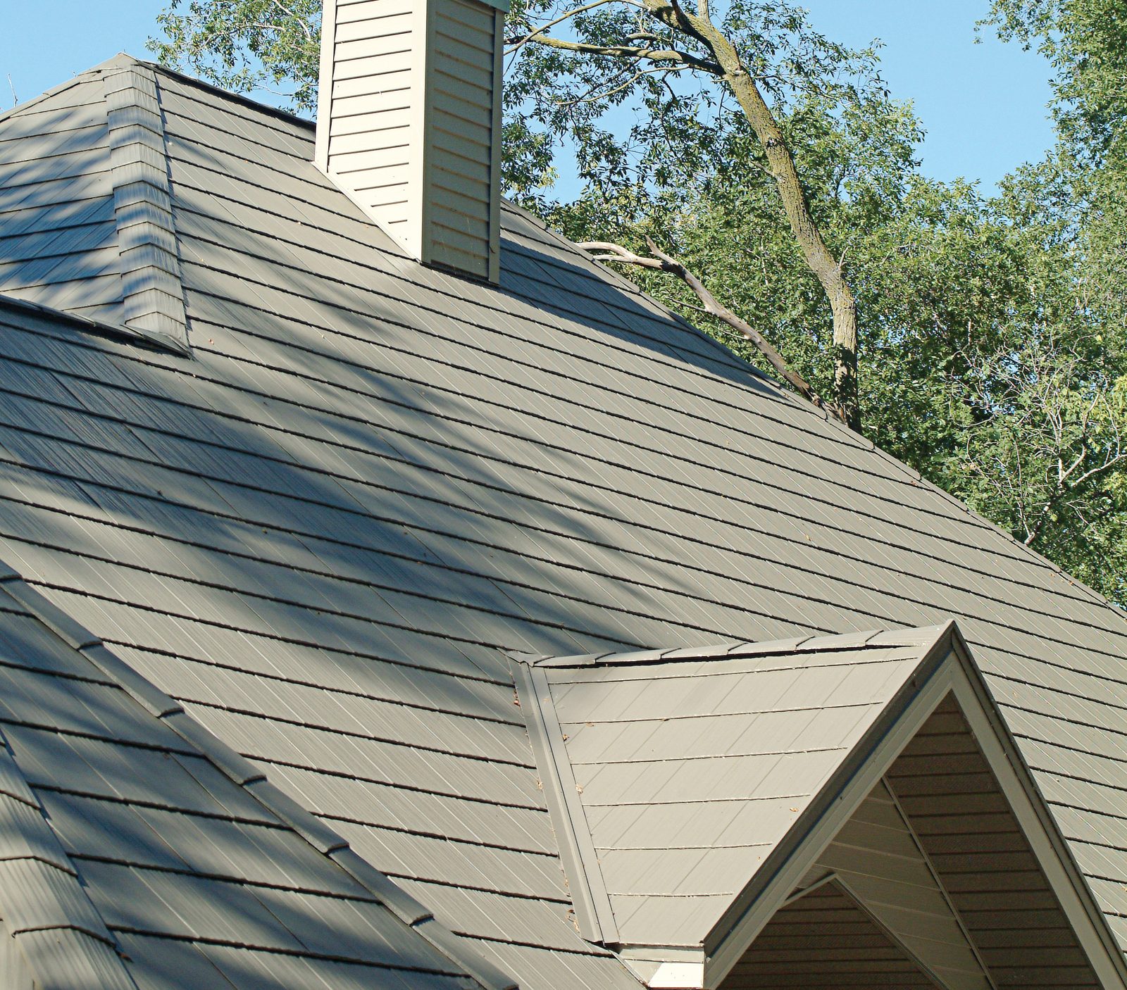 Olive-gray steel shake roofing runs across intersecting roof planes past a sided chimney chase.