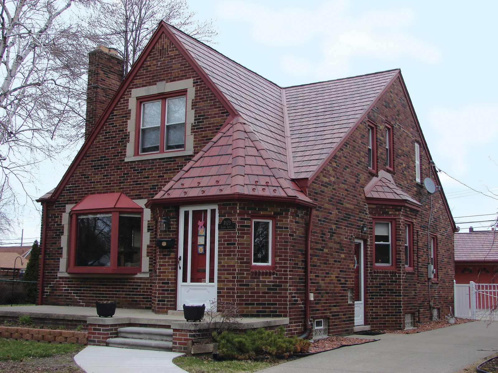 Red-toned slate-look steel roofing covers the steep gables and entry turret of a brick cottage.