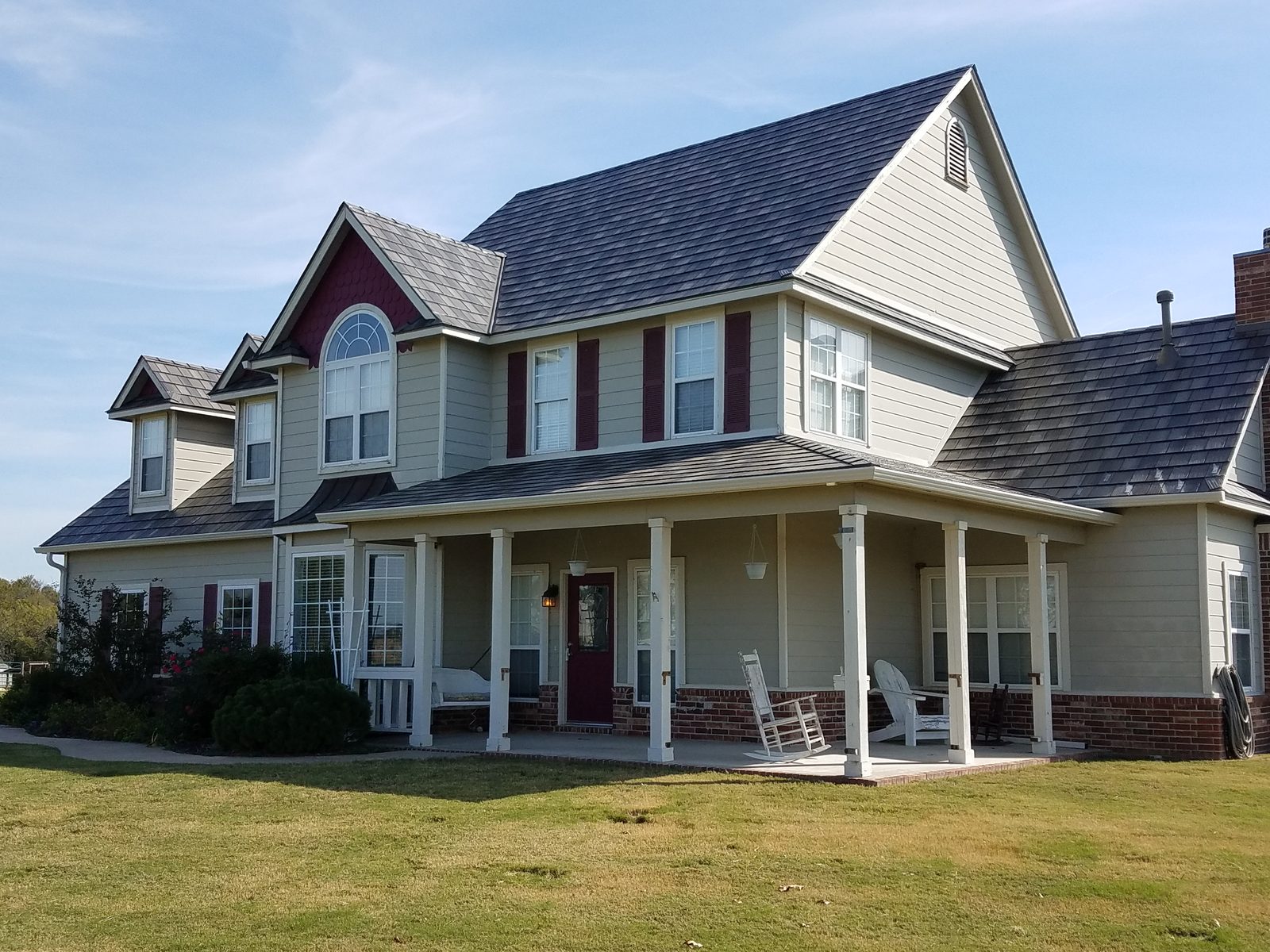 A two-story home under gray steel shake roofing, with a brick-skirted porch and arched gable window.