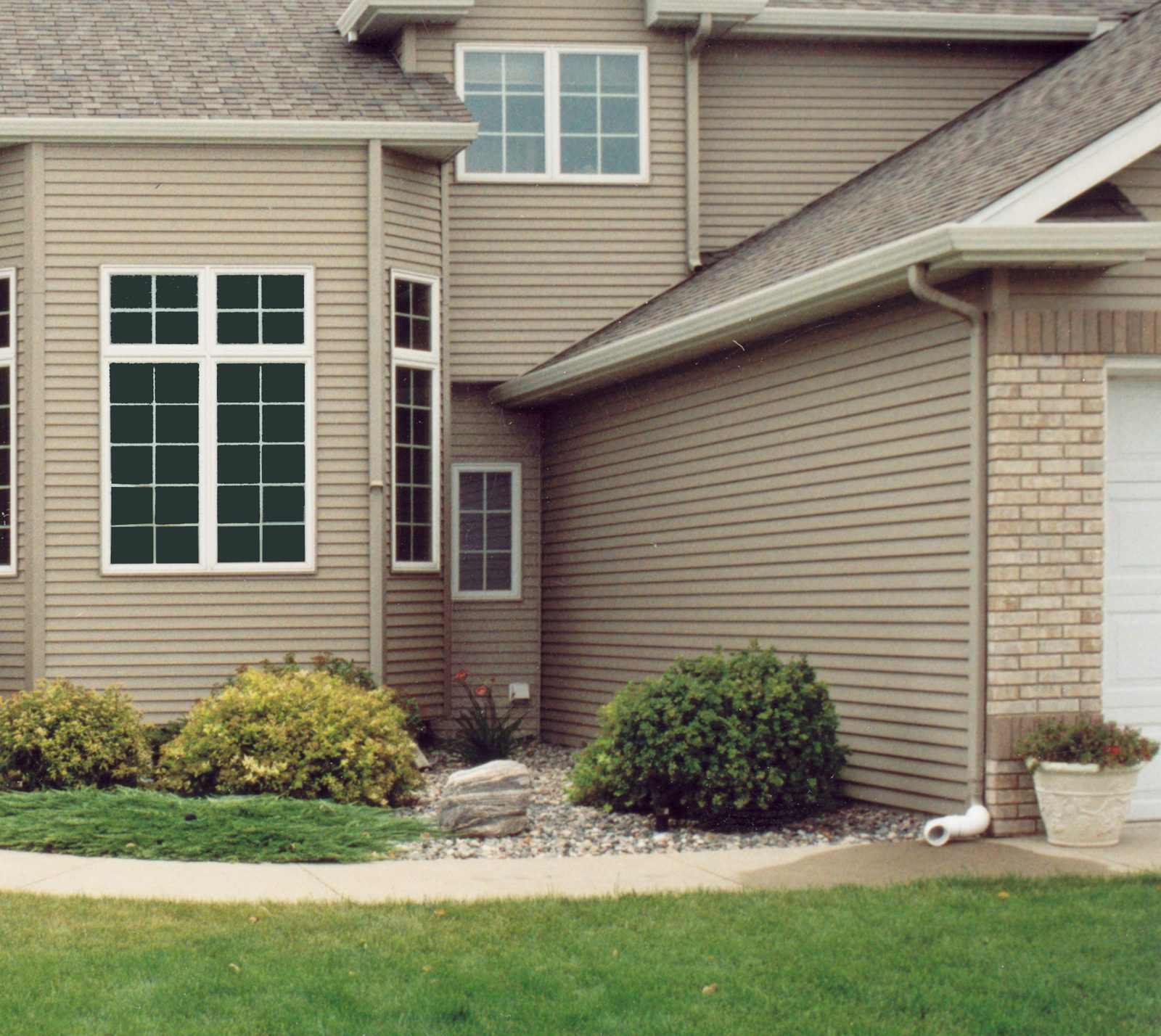 Tan lap siding wraps the corner of a two-story home beside a brick garage wall and foundation beds.