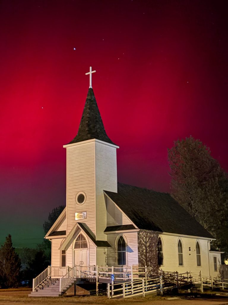 White lap siding and a dark steeple on a small country church beneath a deep red night sky.