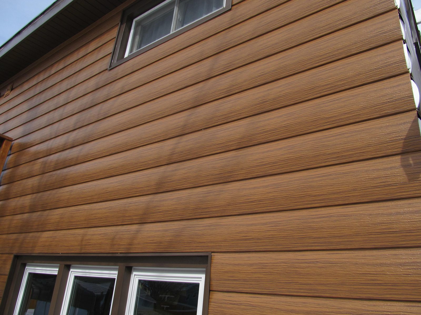 Wood-look lap steel siding runs across a sunlit wall past a small brown-trimmed window.