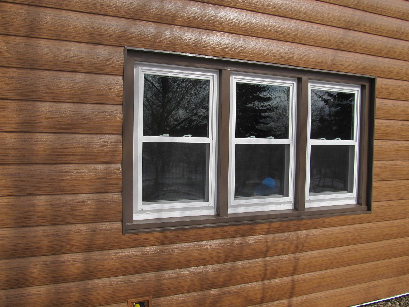 Wood-look steel lap siding surrounds a triple window with brown trim and white sashes.