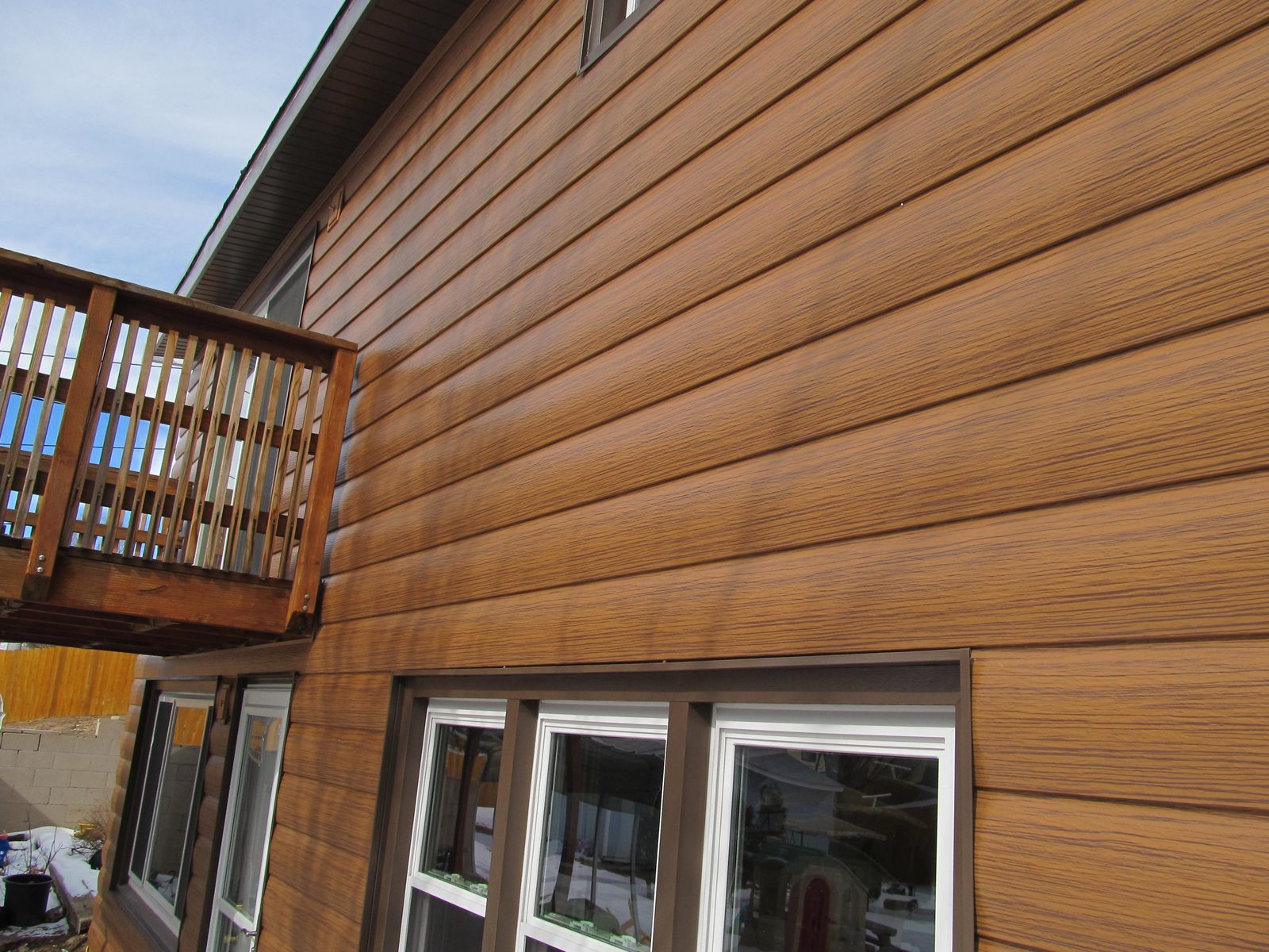 Wood-look steel lap siding covers a two-story wall beside a weathered wood deck and railing.