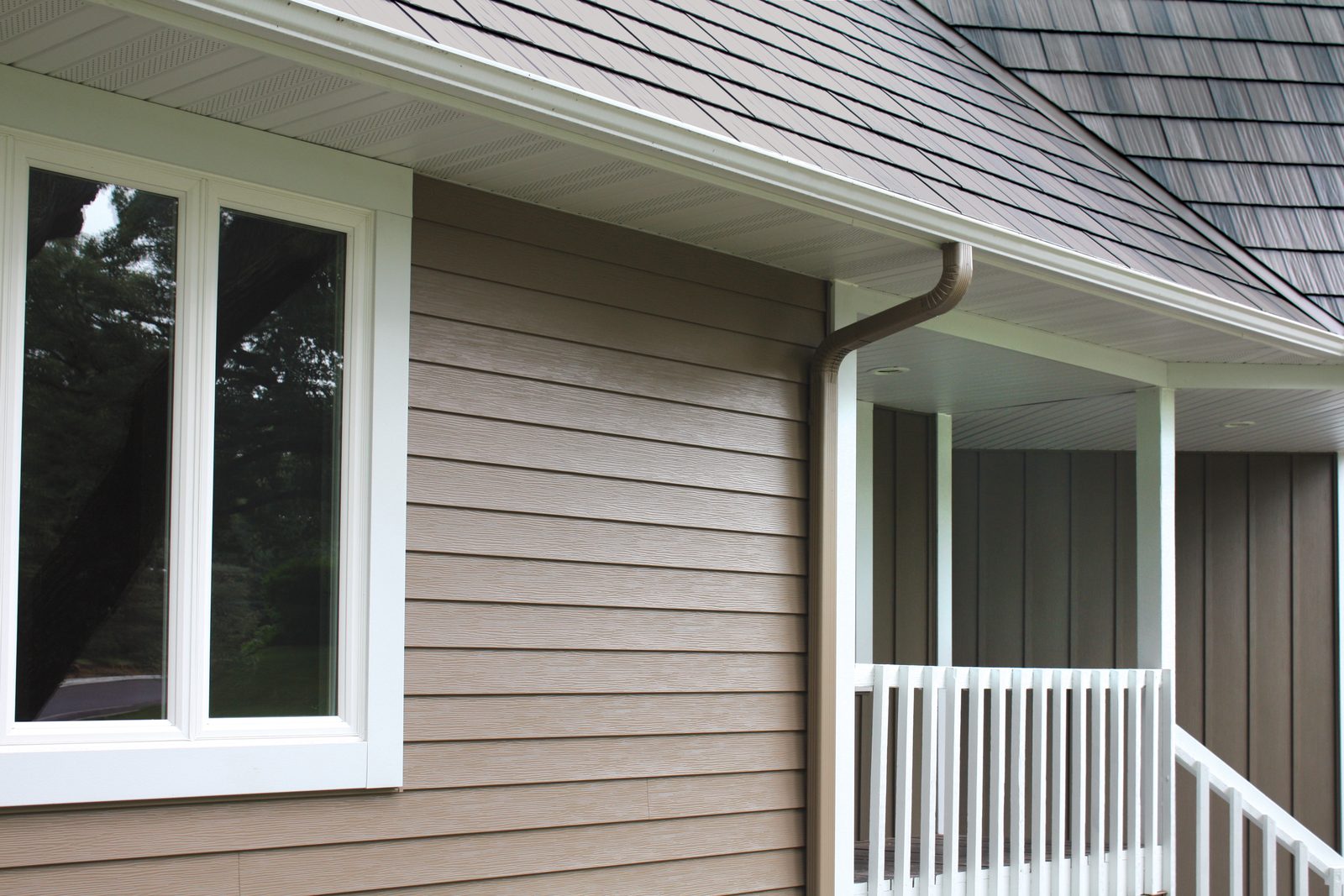 Tan lap steel siding meets a white soffit and downspout below a gray shake roof and porch rail.