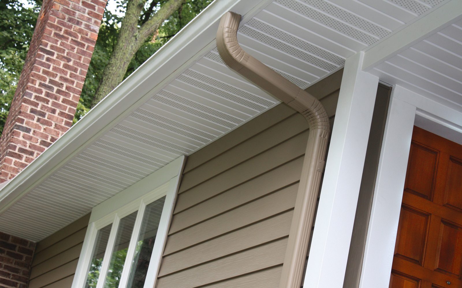 Sage lap steel siding meets a beaded white soffit and a tan downspout near a wood entry door.