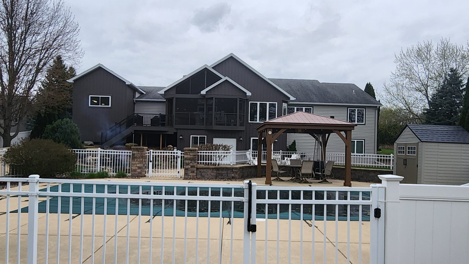 Dark board-and-batten and lap steel siding face a rear elevation behind a fenced pool and gazebo.