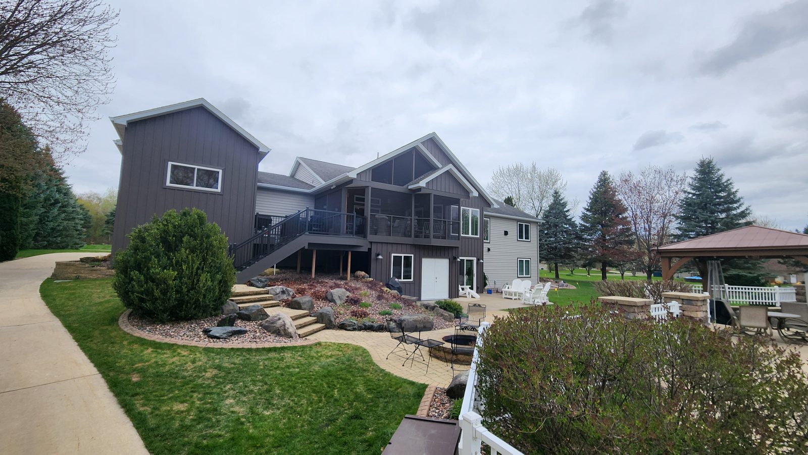 Wide rear yard view of a home in dark board-and-batten and pale lap steel siding above a stone terrace.
