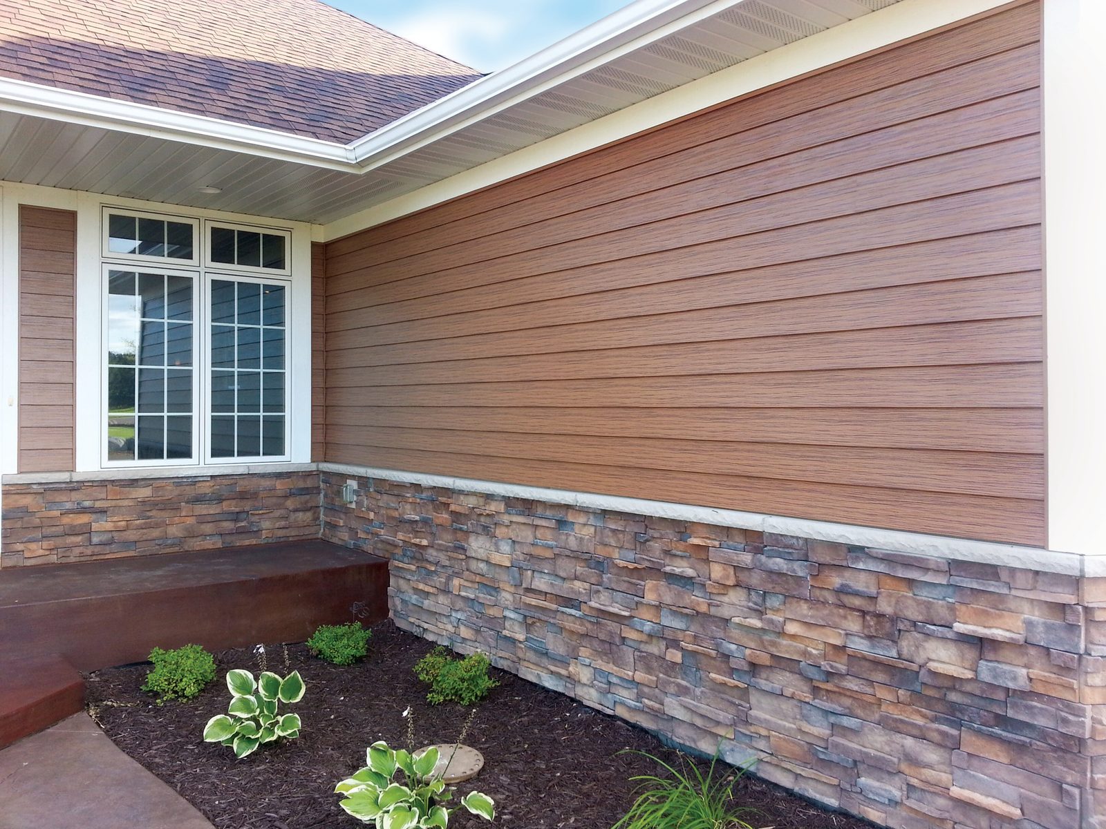 Wood-look lap steel siding runs above a layered stone wainscot beside a white-trimmed bay window.